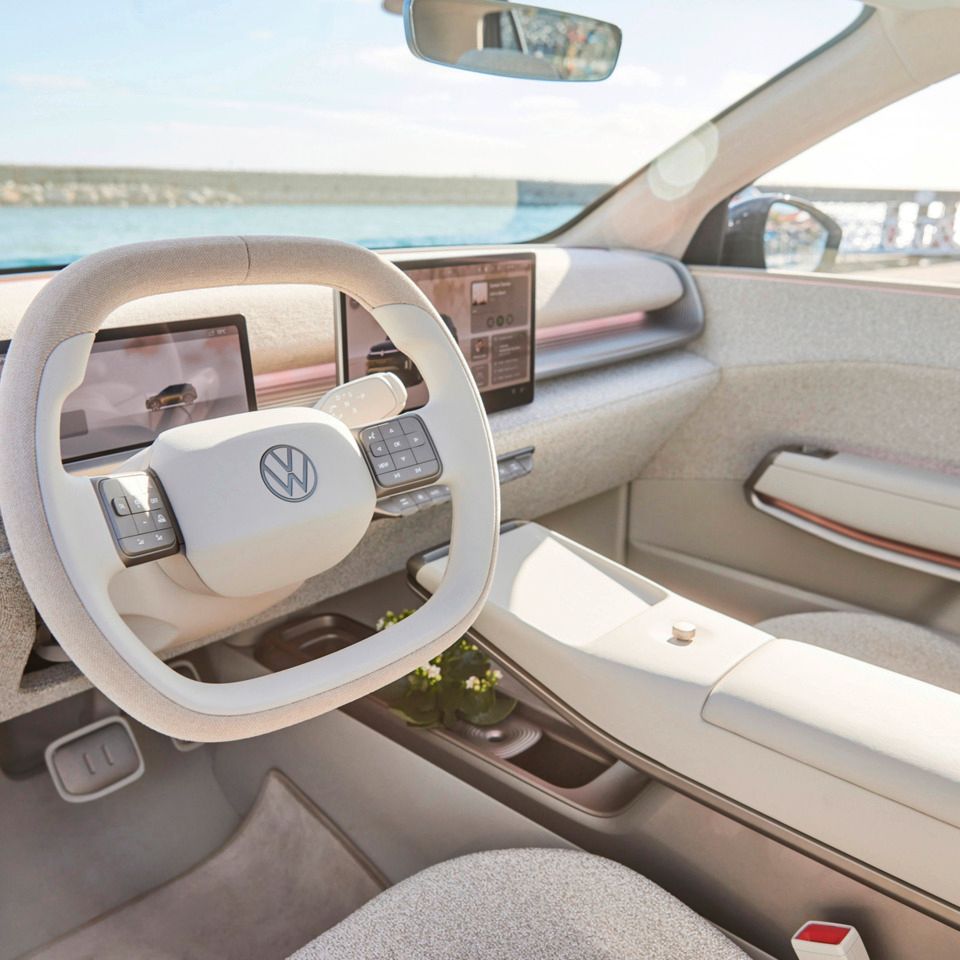 Slika prikazuje notranjost ID. Cross z svetlo sivim oblazinjenjem. Volan je nameščen na sredini, na armaturni plošči pa je več digitalnih zaslonov. V ozadju je skozi vetrobransko steklo viden pogled na vodo.