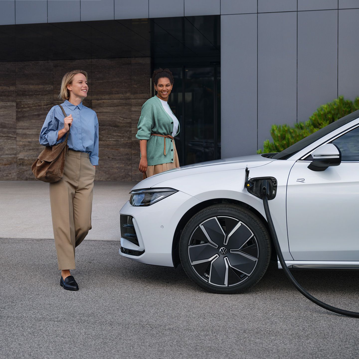 Pogled od strani na bel VW Passat s priključenim polnilnim kablom pred poslovno stavbo; vozilu se približujeta dve ženski.