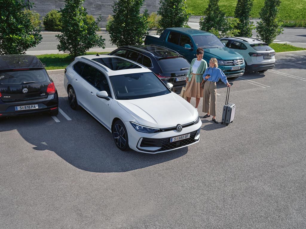 Dve ženski ob belem VW Passatu, ki samodejno parkira s pomočjo opcijskega sistema Park Assist Plus. Ena od žensk drži v roki pametni telefon.