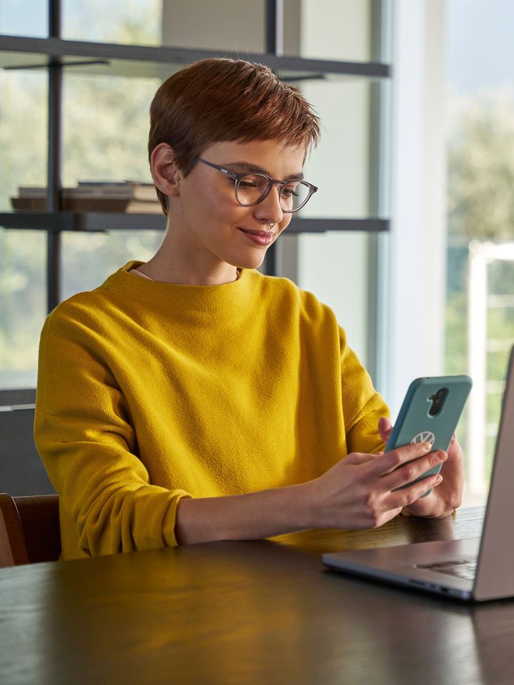Frau schaut vor ihrem Laptop auf Ihr Handy und prüft, wo ihr Volkswagen gerade ist.