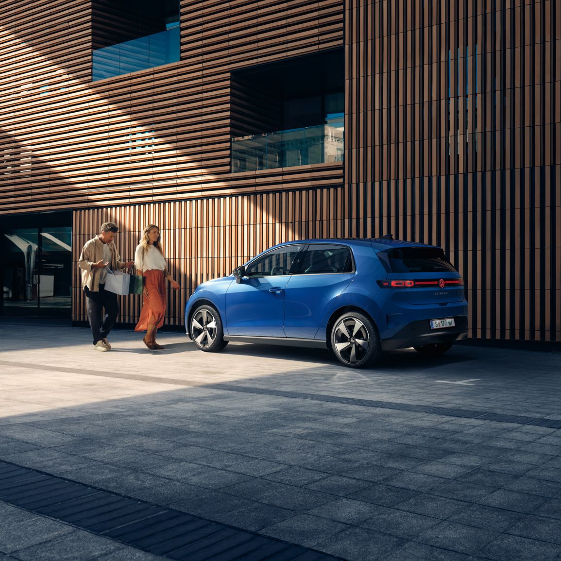 Modri VW ID. Polo Style stoji pred stavbo z leseno fasado. Moški in ženska z nakupovalnimi vrečkami hodita proti njej