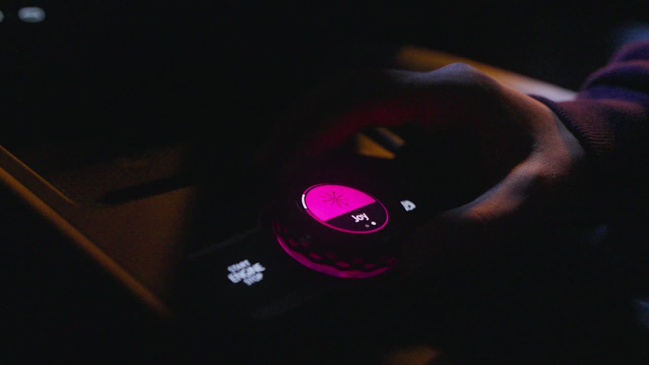 Oseba upravlja osvetljen gumb v vozilu. Gumb je osvetljene rozne barve in je videti v temi. 
