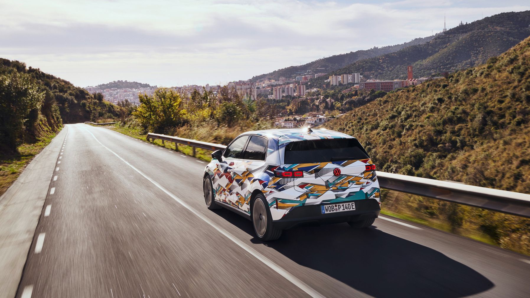 Zakamufliran ID. Polo vozi po hriboviti cesti, pogled od zadaj. V ozadju je mesto, obdano z naravo in hribi. 