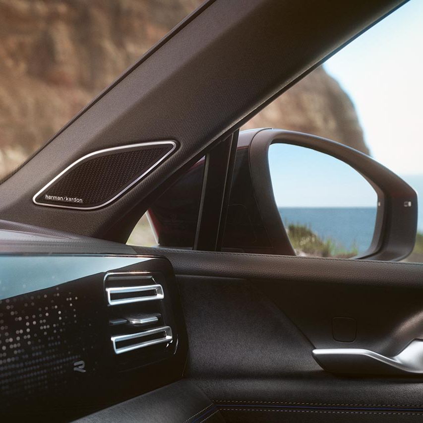 Notranjost sodobnega avtomobila VW Golf Variant s pogledom na vrata in armaturno ploščo, kjer izstopa zvočnik Harman Kardon v A-stebričku. Na levi je del digitalne armaturne plošče z dekorativnim vzorcem, na desni pa stransko ogledalo in okno, skozi katera se vidi skalnata stena in morje v ozadju.