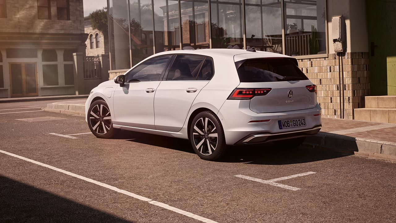 Bel avtomobil Volkswagen Golf je parkiran ob pločniku v mirni mestni ulici. Vozilo je belo, petvratno, s sodobnimi LED zadnjimi lučmi in temnimi stekli. V ozadju so steklene fasade in starejše stavbe v toplih sončnih tonih.
