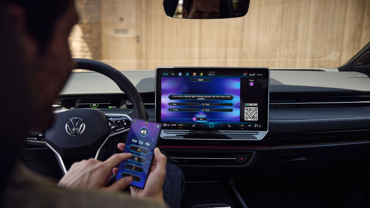 Moški na voznikovem sedežu igra igro »Kdo želi postati milijonar?« s pametnim telefonom kot krmilnikom. na zaslonu VW infotainment sistema