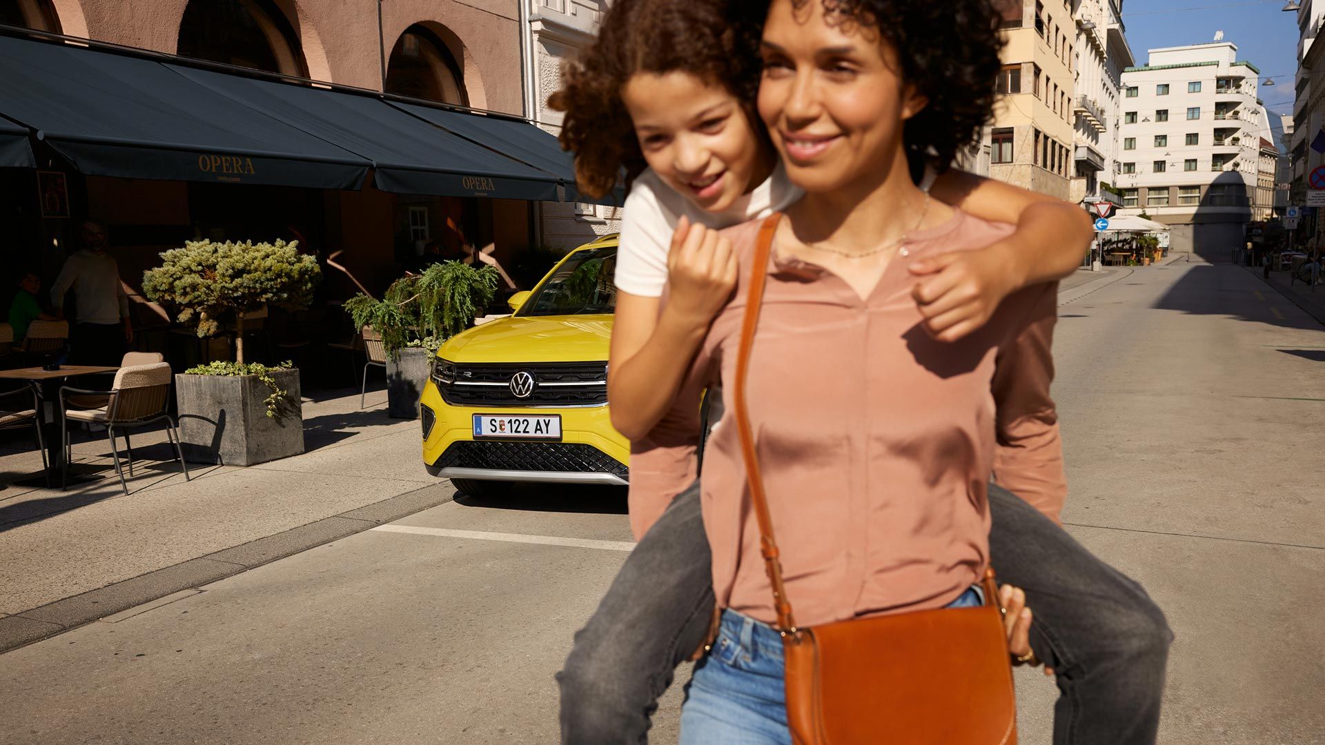 Na ulici v mestnem središču ženska na hrbtu nosi deklico. V ozadju je viden rumen VW T-Cross.