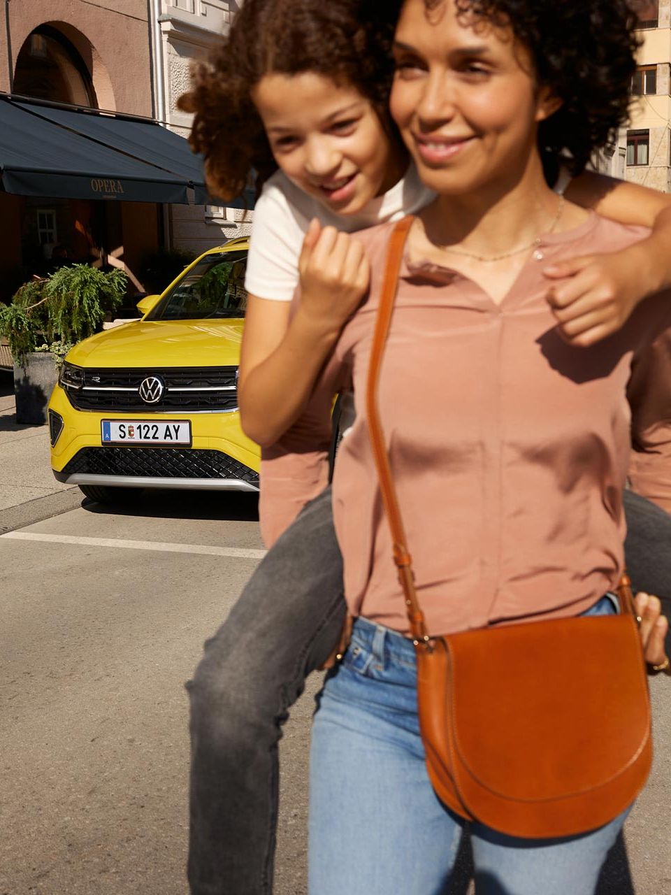 Na ulici v mestnem središču ženska na hrbtu nosi deklico. V ozadju je viden rumen VW T-Cross.