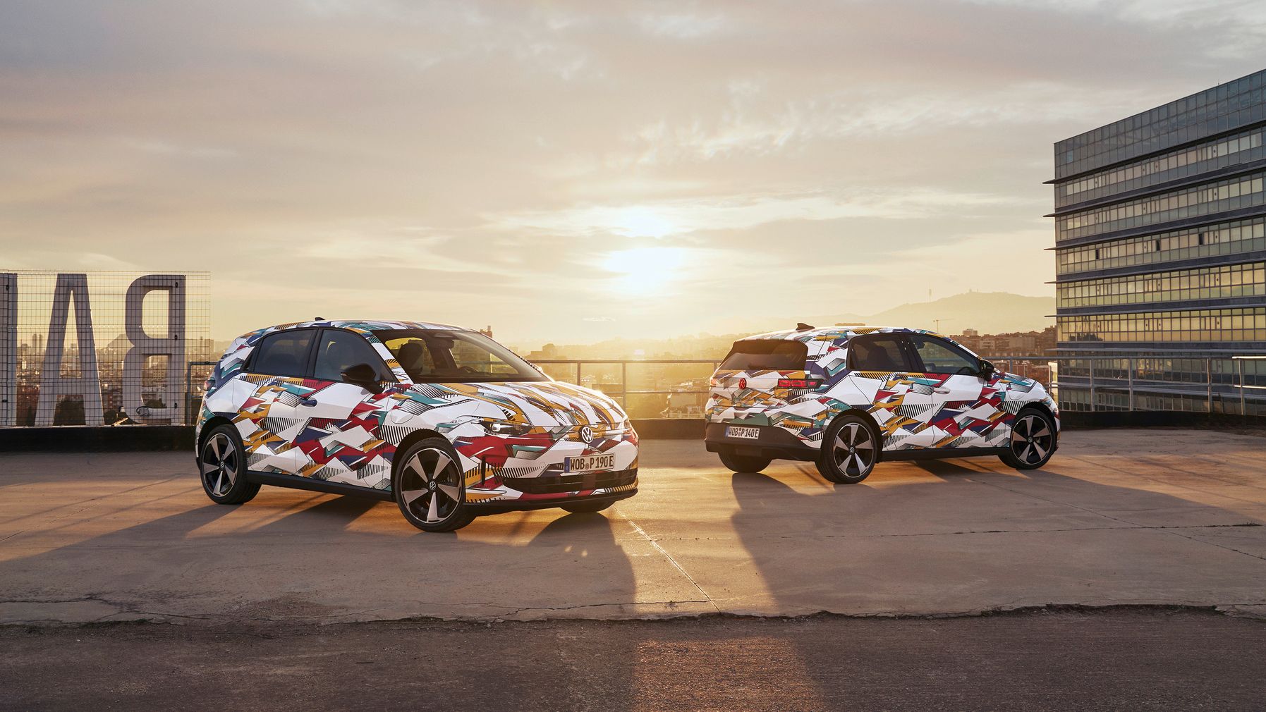 Na sliki sta dva zakamuflirana modela ID. Polo, parkirana na strehi zgradbe ob sončnem zahodu. V ozadju je vidna moderna steklena zgradba. 