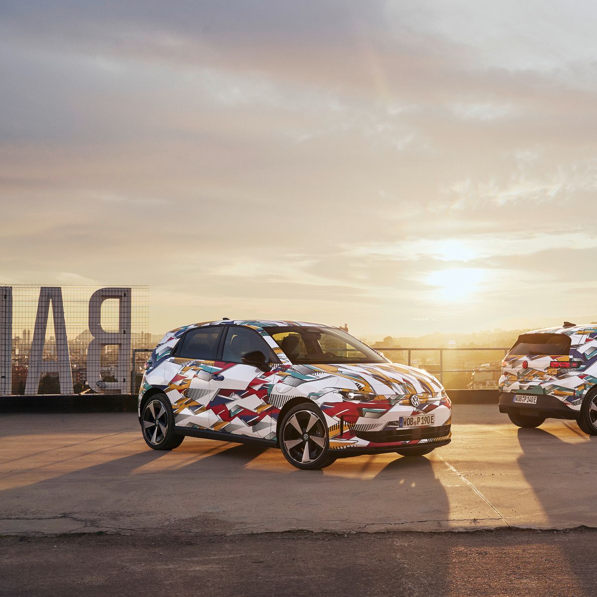 Na sliki sta dva zakamuflirana modela ID. Polo, parkirana na strehi zgradbe ob sončnem zahodu. V ozadju je vidna moderna steklena zgradba. 