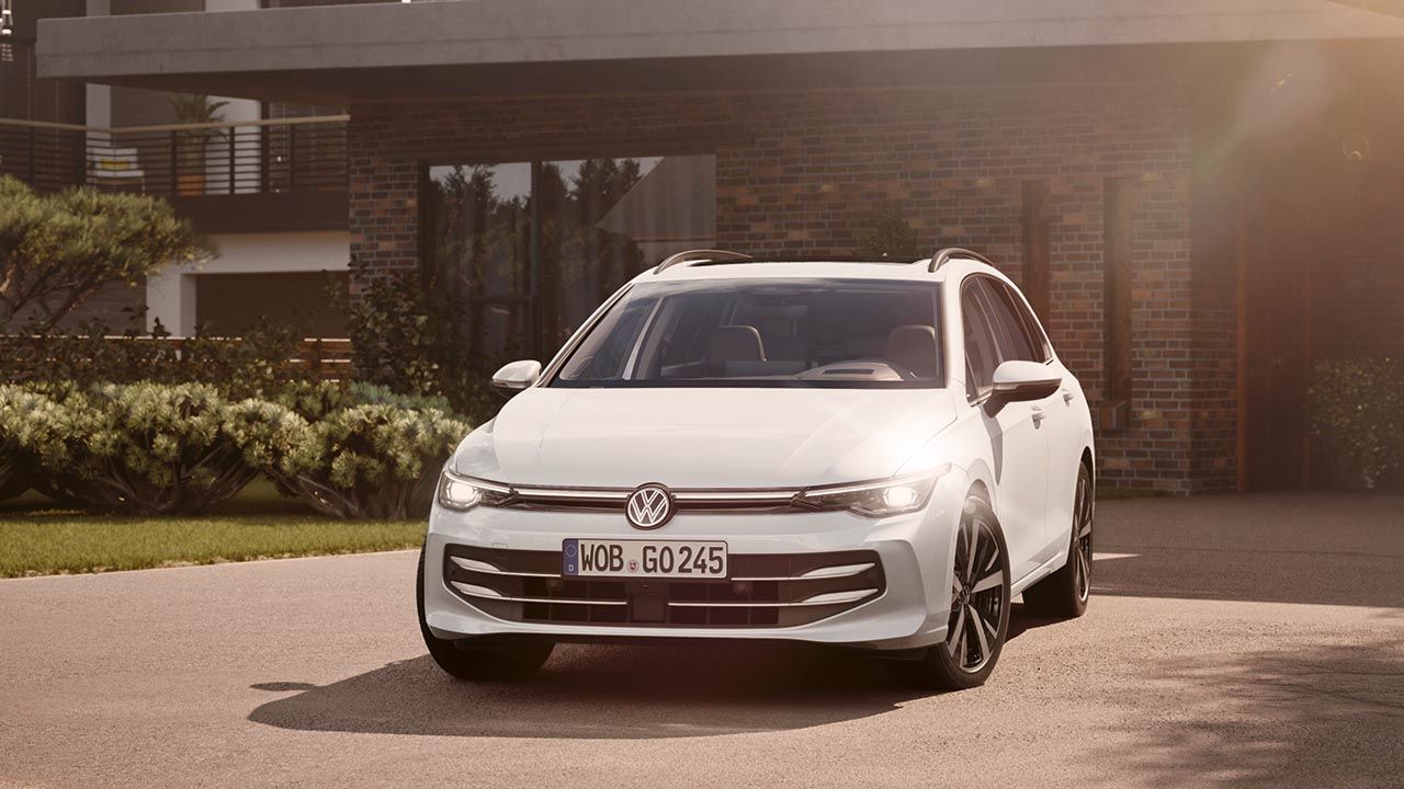 Bel VW Golf Variant je parkiran na dovozni poti pred moderno opečnato hišo z velikimi okni. Sončna svetloba osvetljuje sprednji del vozila in ustvarja mehko, toplo vzdušje. V ozadju so urejene zelenice in grmičevje, ki poudarjajo eleganten videz avtomobila.