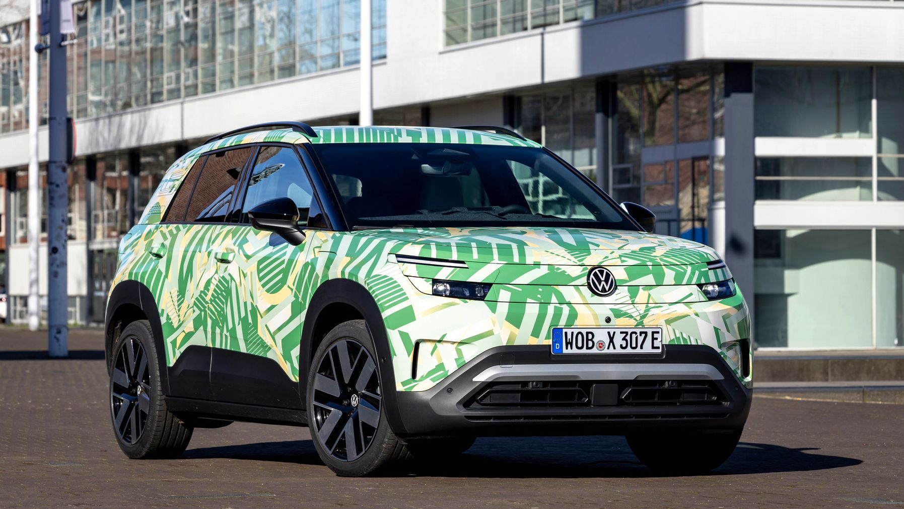 Pred sodobnimi stavbami stoji električni SUV Volkswagen. Barvito vozilo ima zeleno-kremno kamuflažno vzorec z motivi listov. 