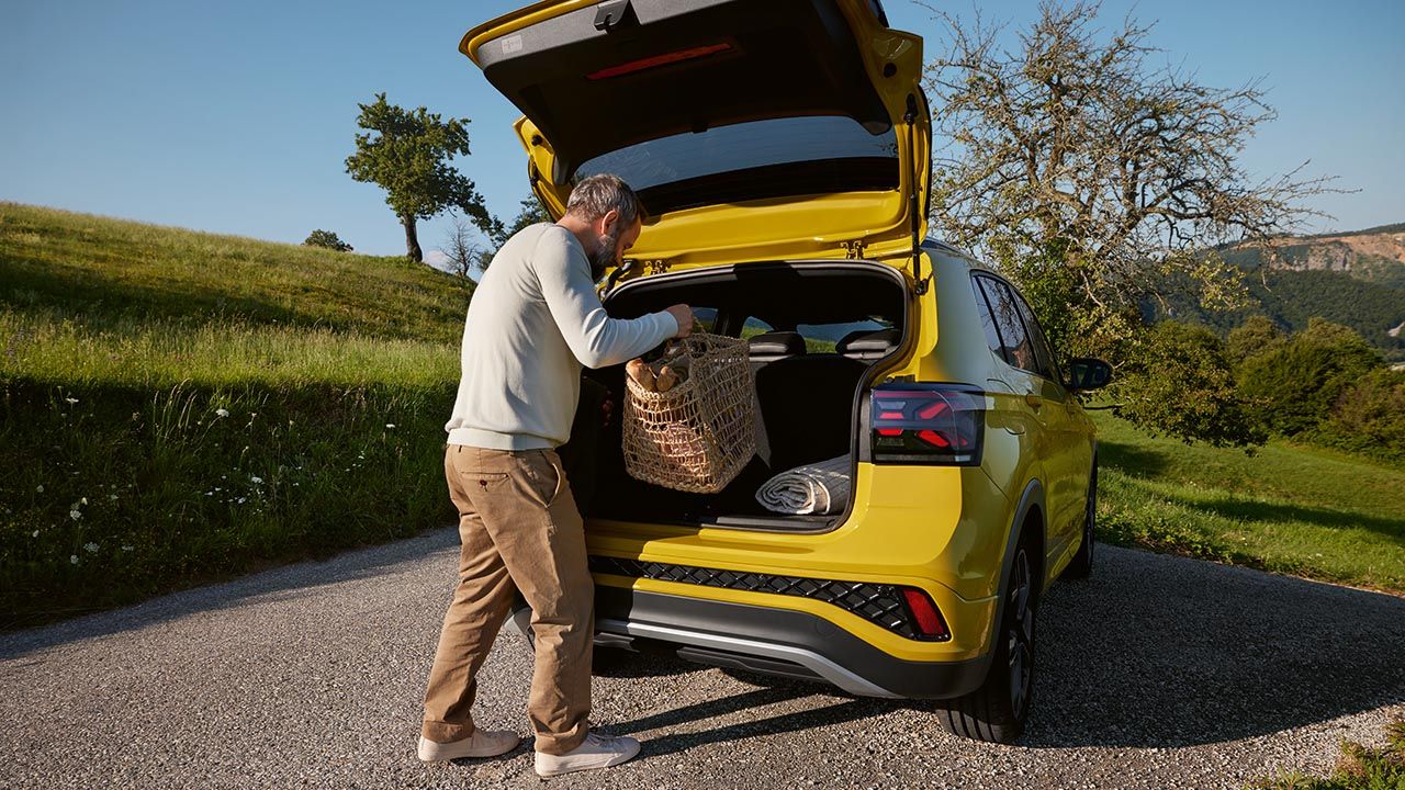 Moški odlaga veliko košaro za piknik v prtljažnik rumenega VW T-Crossa, ki stoji na cesti sredi hribovite pokrajine.