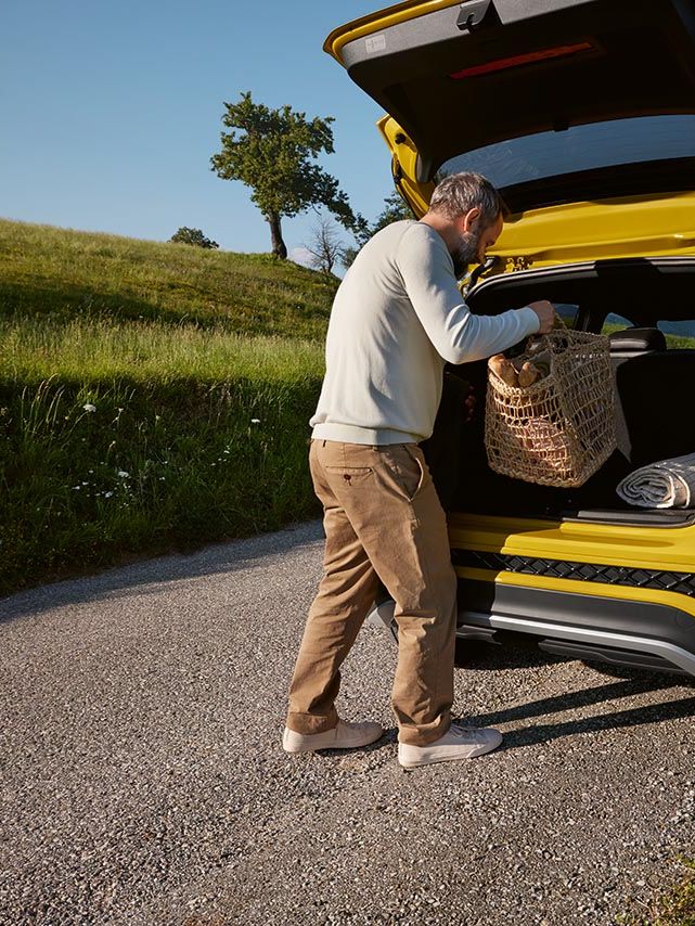 Moški odlaga veliko košaro za piknik v prtljažnik rumenega VW T-Crossa, ki stoji na cesti sredi hribovite pokrajine.