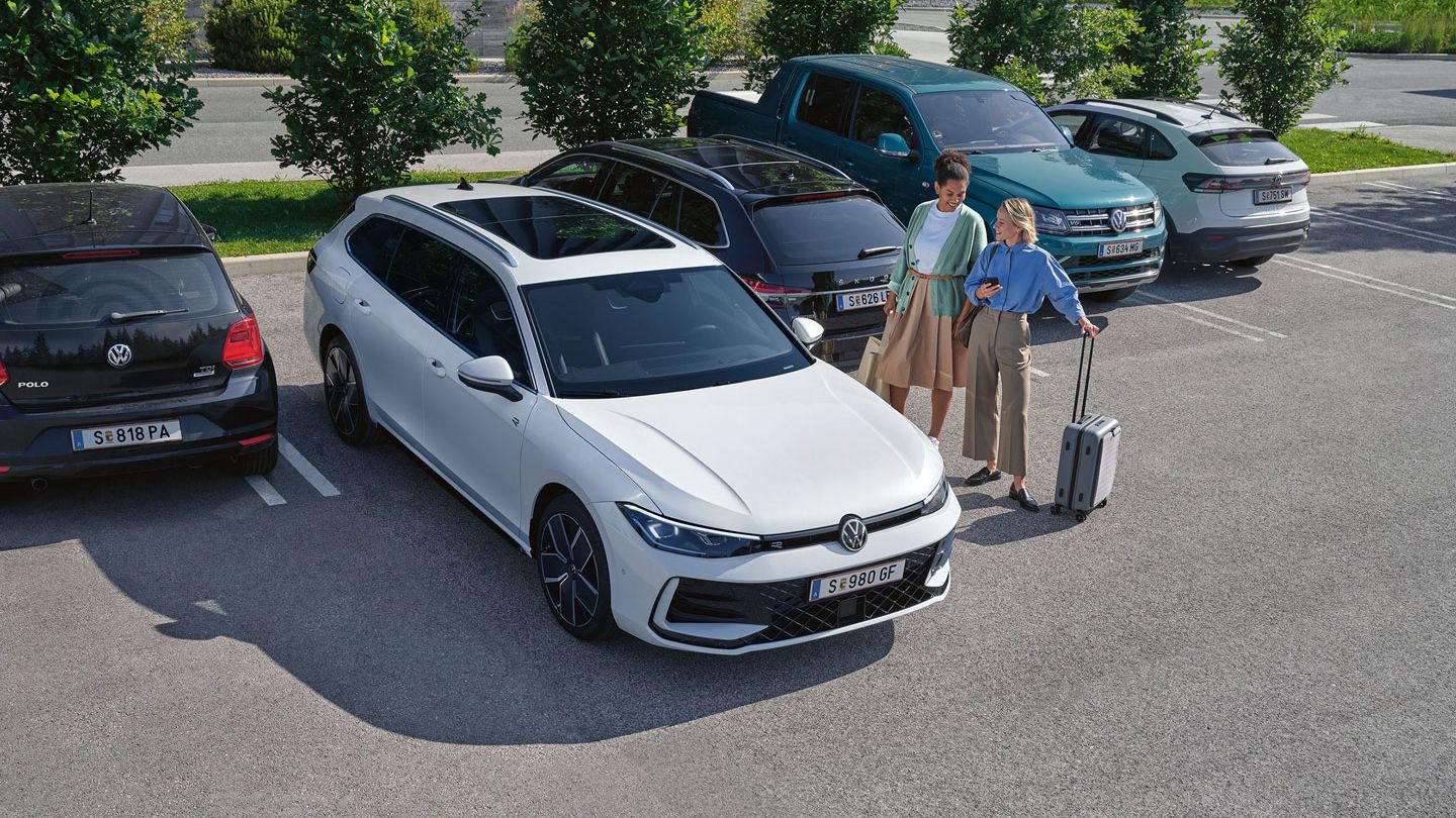 Dve osebi stojita na parkirišču poleg belega avtomobila Volkswagen. Ena oseba je oblečena v modro srajco in drži majhen kovček. Za avtomobilom je parkiranih več drugih vozil.