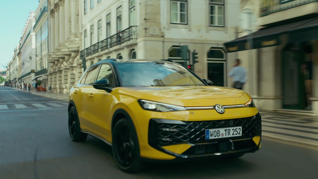 Rumeni Volkswagen T-Roc vozi po mestni ulici, obdani z starimi stavbami. Ljudje so vidni v ozadju na ulici.