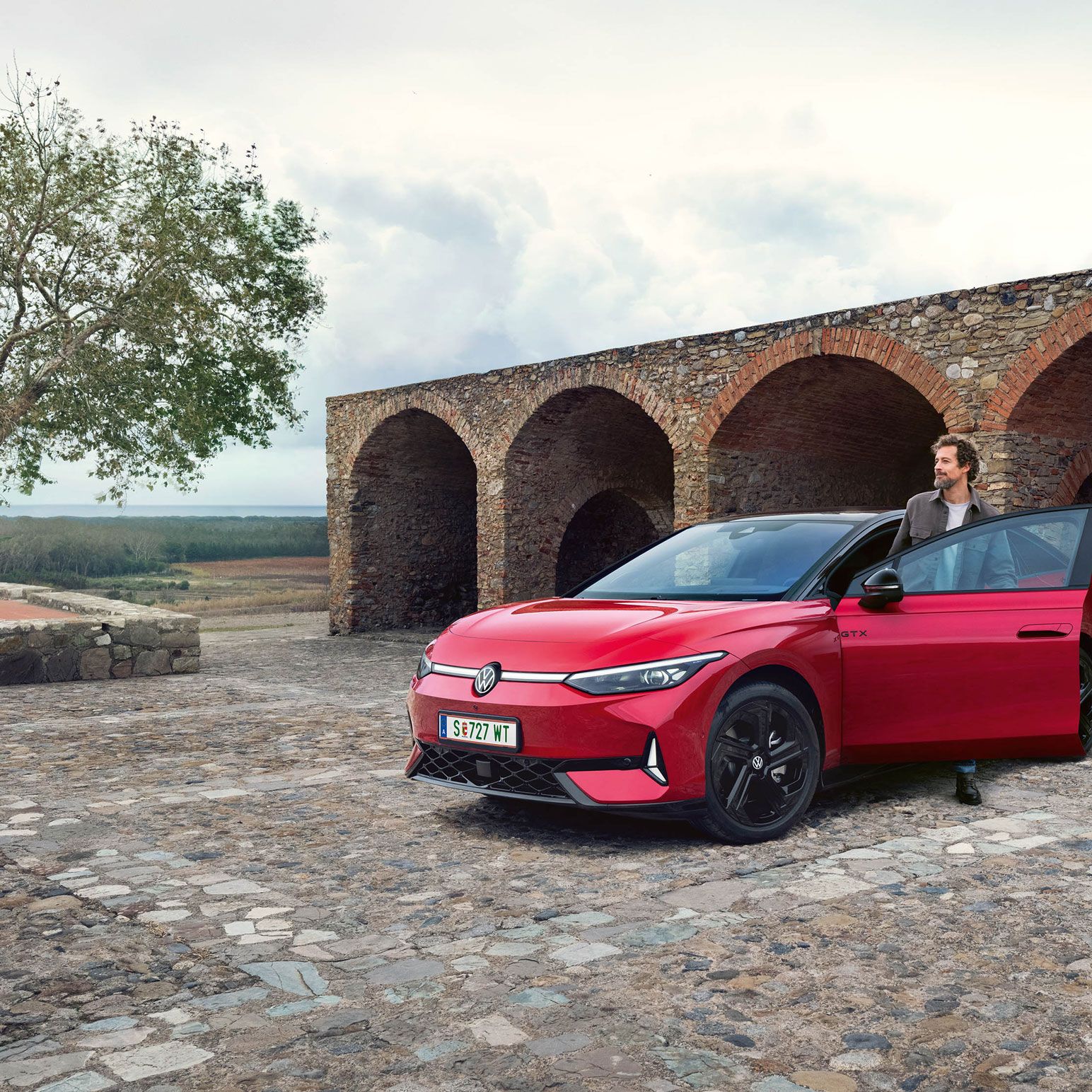 Rdeč VW ID.7 GTX parkiran pred kamnito zgradbo s tremi oboki. Oseba stoji ob odprtih vratih voznika. 