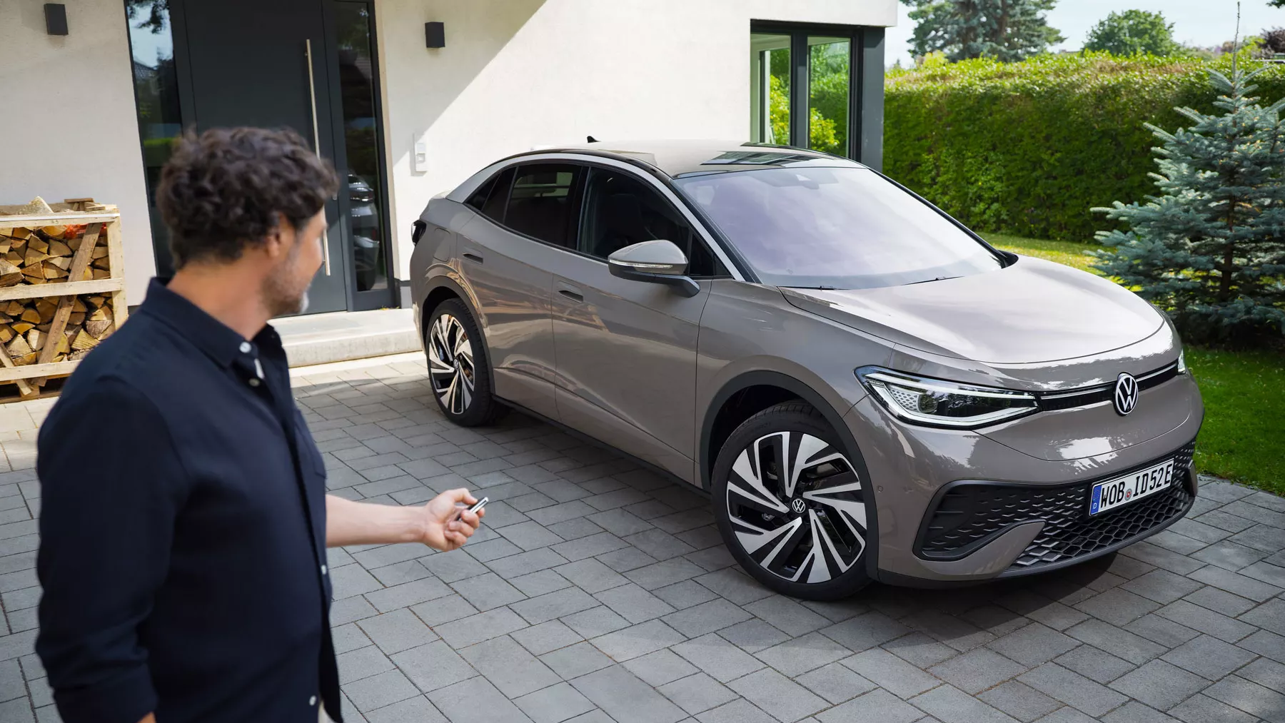Moški stoji ob levem robu slike in gleda nazaj proti svetlo sivemu VW ID.5, ki je parkiran na dovozu pred hišo. Moški ima radijski daljinski upravljalnik in ga drži usmerjenega proti avtomobilu.