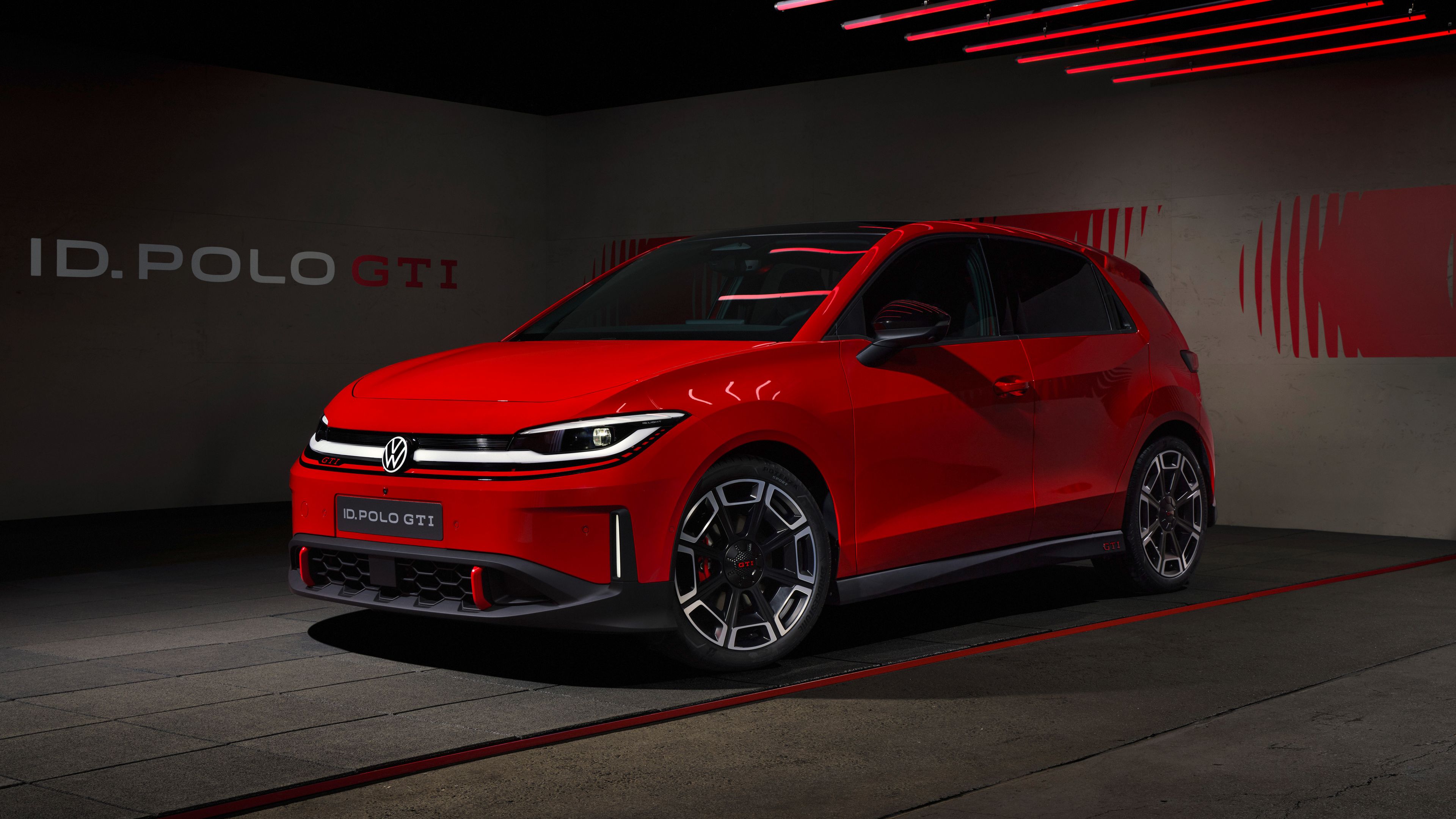 Rdeč športni Volkswagen ID. Polo GTI je parkiran v temnem, modernem studiu z rdečimi LED-lučmi na stropu. Avto ima agresiven sprednji del z ozko svetlobno linijo, velikimi športnimi platišči in nizkim profilom. V ozadju je na steni napis “ID. POLO GTI” in grafični rdeči motiv.
