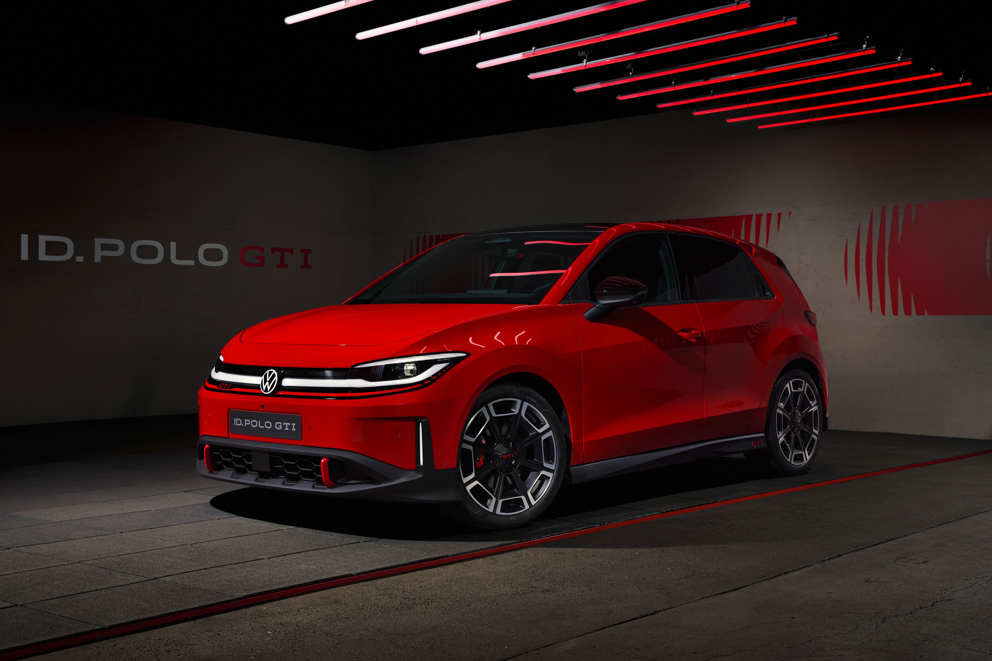 Rdeč športni Volkswagen ID. Polo GTI je parkiran v temnem, modernem studiu z rdečimi LED-lučmi na stropu. Avto ima agresiven sprednji del z ozko svetlobno linijo, velikimi športnimi platišči in nizkim profilom. V ozadju je na steni napis “ID. POLO GTI” in grafični rdeči motiv.