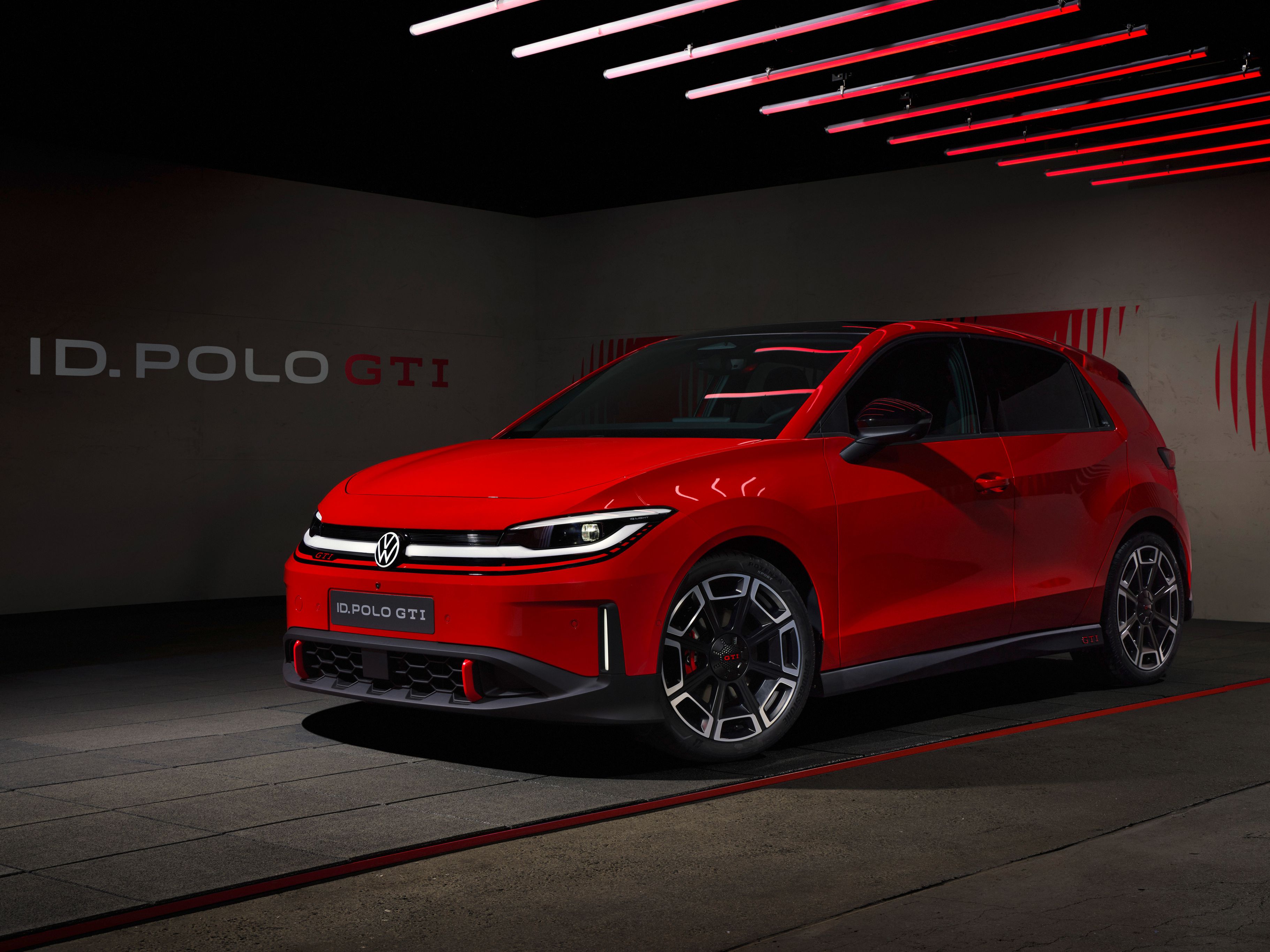 Rdeč športni Volkswagen ID. Polo GTI je parkiran v temnem, modernem studiu z rdečimi LED-lučmi na stropu. Avto ima agresiven sprednji del z ozko svetlobno linijo, velikimi športnimi platišči in nizkim profilom. V ozadju je na steni napis “ID. POLO GTI” in grafični rdeči motiv.