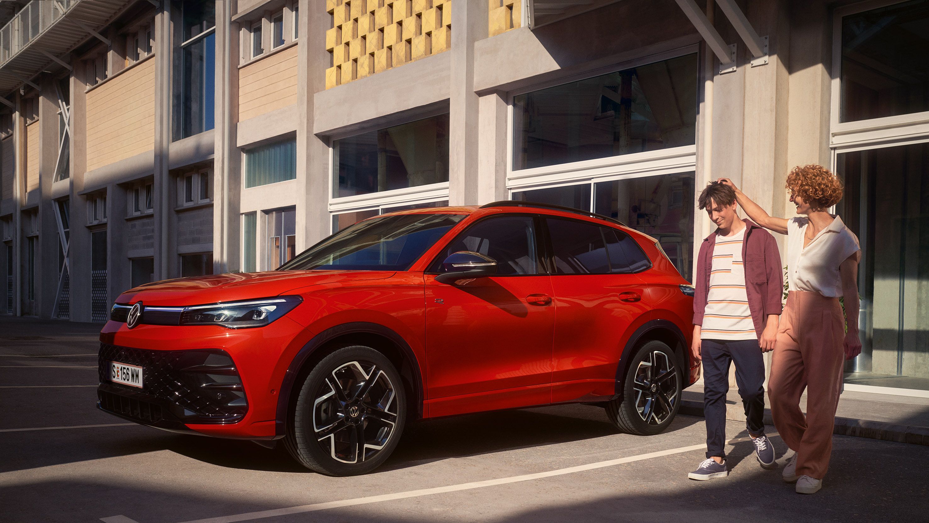 Rdeč VW Tiguan je parkiran pred stavbo, mimo gresta ženska in mlad fant.