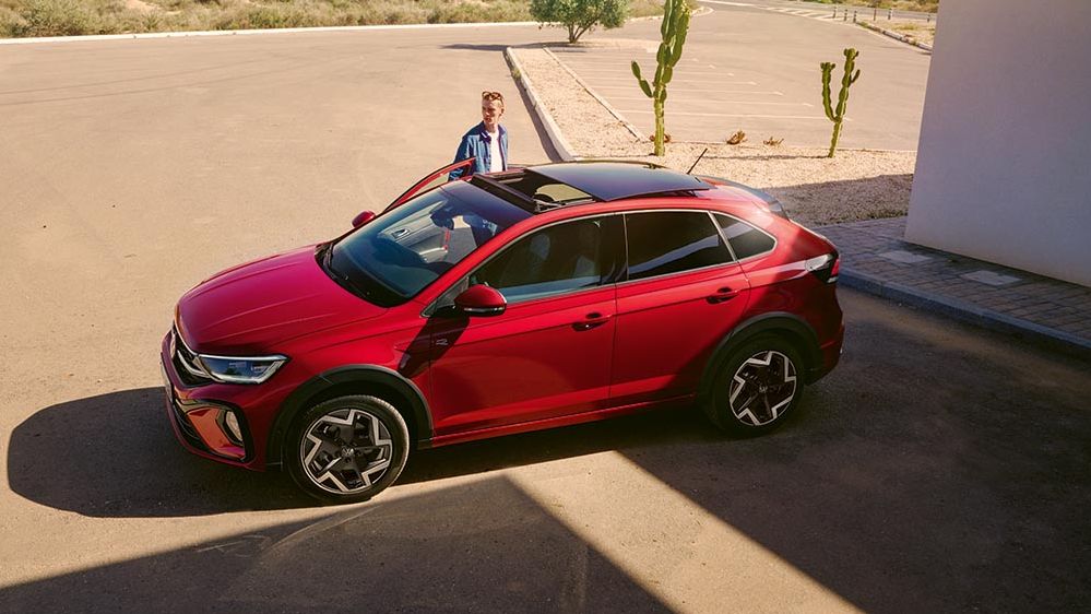 Rdeč športni Volkswagen Taigo je parkiran na sončnem, praznem parkirišču v puščavski pokrajini. Ob avtomobilu stoji moški v srajci in gleda proti stavbi z belimi stenami na desni strani slike. V ozadju so nizka grmičevja, kaktusi in hribovita krajina pod modrim nebom.