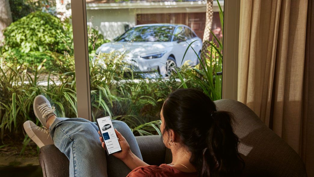 Ženska s pametnim telefonom v roki sproščeno sedi v dnevni sobi in skozi okno gleda VW ID.7.