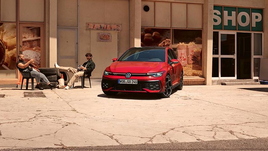 Volkswagen Golf GTI je parkiran pred majhno trgovino z napisom »SHOP« na pročelju. Na levi strani pred trgovino sedita dve osebi na klopi, ena leži z nogami na drugi klopi. Okolica deluje suha in sončna, z razpokanimi tlemi na parkirišču.