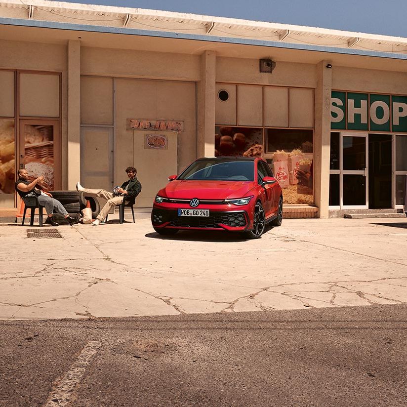 Volkswagen Golf GTI je parkiran pred majhno trgovino z napisom »SHOP« na pročelju. Na levi strani pred trgovino sedita dve osebi na klopi, ena leži z nogami na drugi klopi. Okolica deluje suha in sončna, z razpokanimi tlemi na parkirišču.