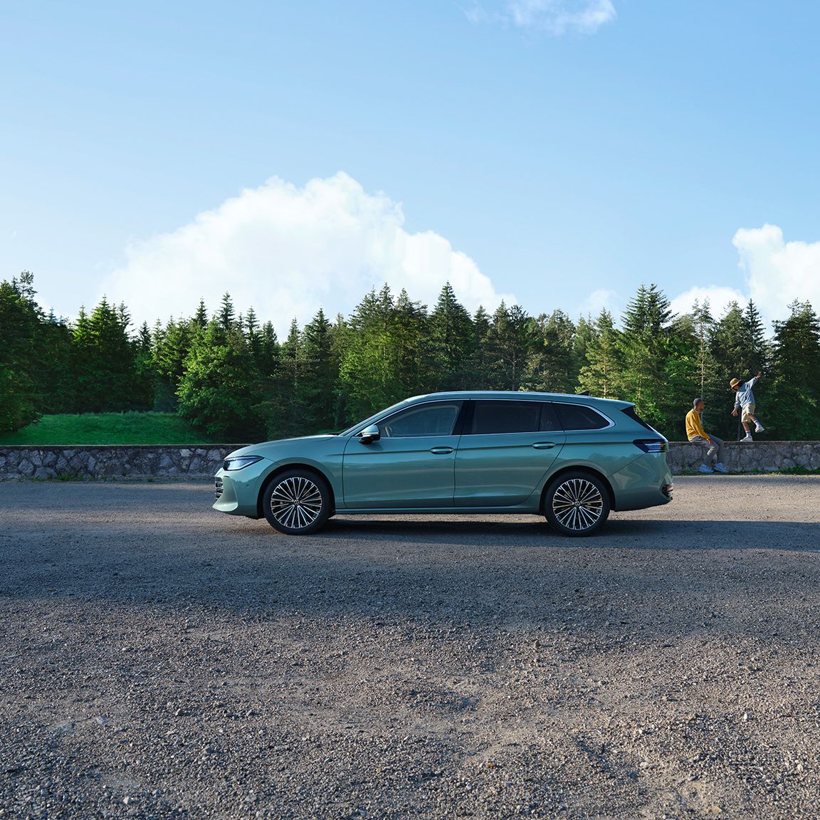 Ein grüner VW Passat B9 in der Seitenansicht, im Hintergrund zwei Menschen und Wald