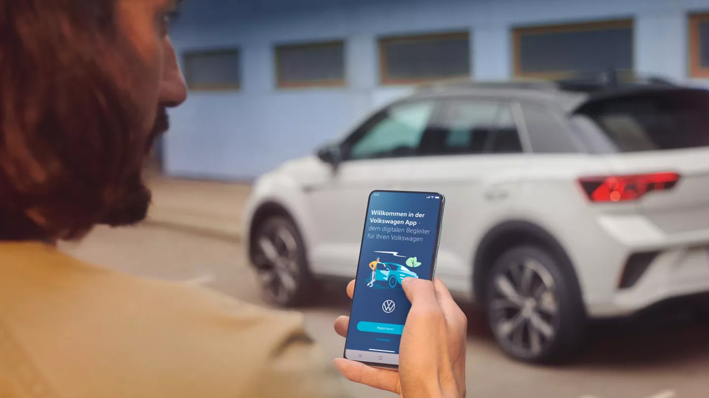 Bel VW T-Roc R-Line pred zgradbo, viden od strani, v ospredju stoji moški s pametnim telefonom.