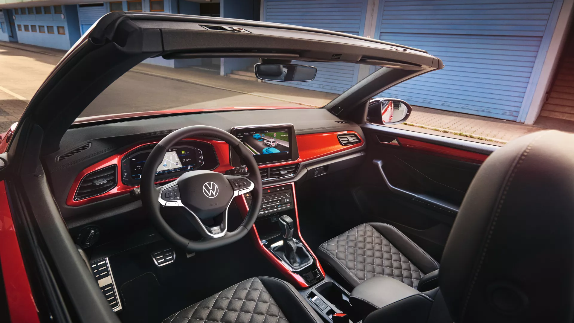 Notranjost modela VW T-Roc Cabriolet: Pogled z zgornje strani skozi odprto streho na armaturno ploščo z rdečimi dekorativnimi elementi in sprednja sedeža. 