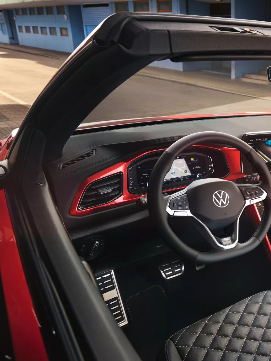 Notranjost modela VW T-Roc Cabriolet: Pogled z zgornje strani skozi odprto streho na armaturno ploščo z rdečimi dekorativnimi elementi in sprednja sedeža. 