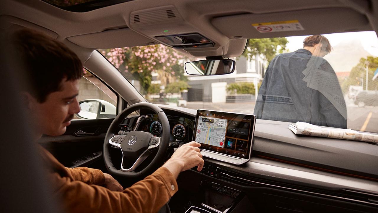 Voznik v Volkswagen Golf sedi za volanom in z dotikom upravlja navigacijo na sredinskem zaslonu. Zunaj vozila stoji druga oseba pri sovoznikovih vratih, na armaturni plošči pa leži zložen časopis. Scena deluje vsakdanja, kot priprava na vožnjo po mestu.