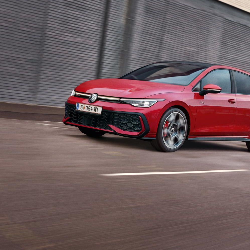 Diagonalno od spredaj je prikazan rdeč VW Golf GTI, ki se pelje mimo sive stavbe.