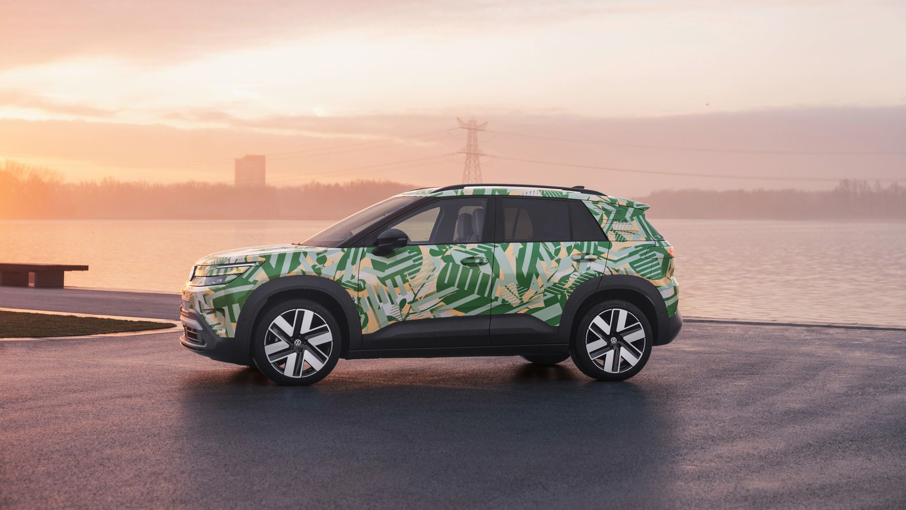 Volkswagen ID. Cross z zeleno-rumenim kamuflažnim vzorcem stoji na asfaltirani površini ob vodi. V ozadju je jezero ali reka z megleno obalo in nežno oranžnim sončnim vzhodom ali zahodom. 