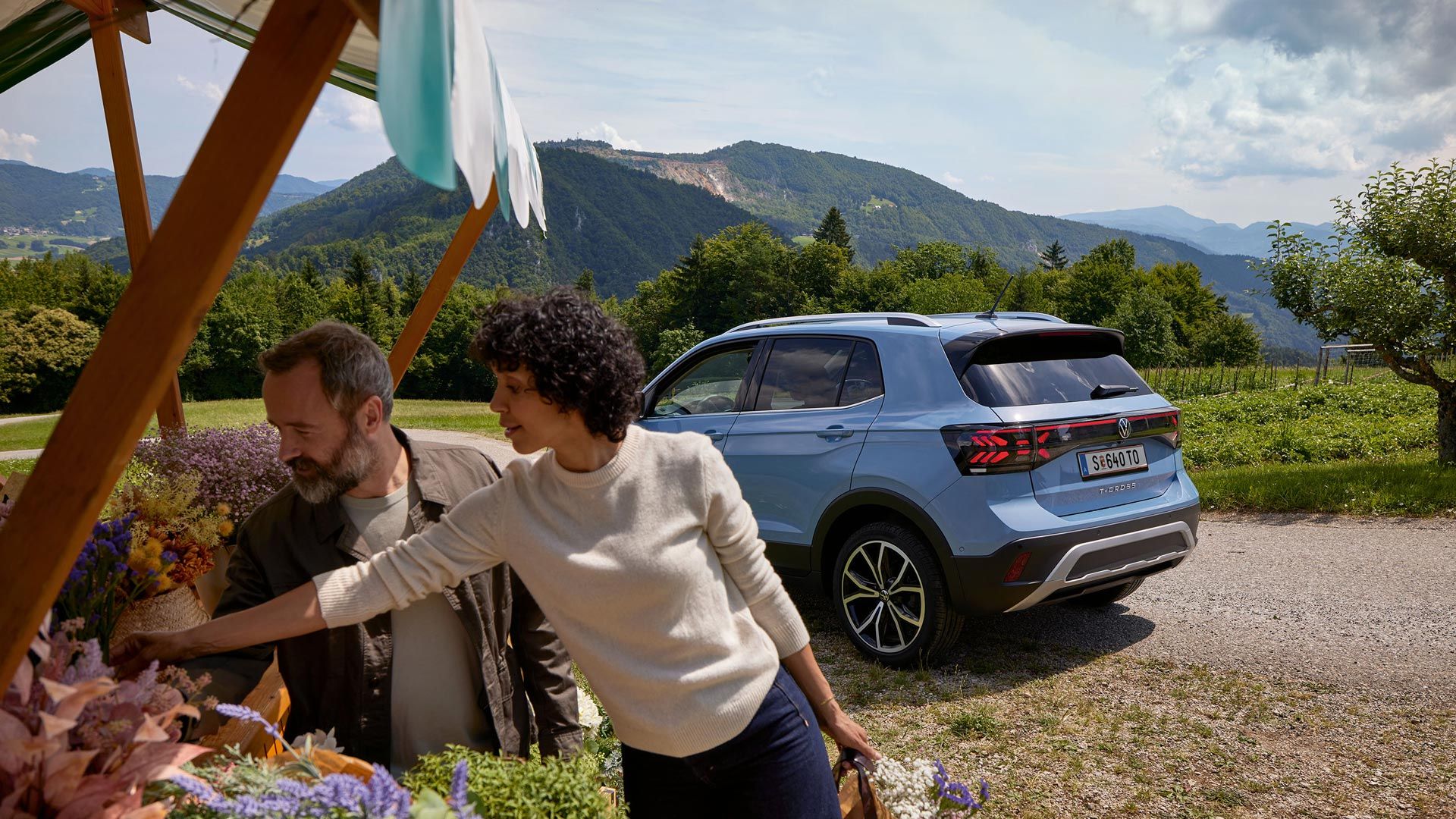 Moški in ženska stojita pred stojnico s cvetjem. Za njima je parkiran moder VW T-Cross, v ozadju je vidno hribovje.