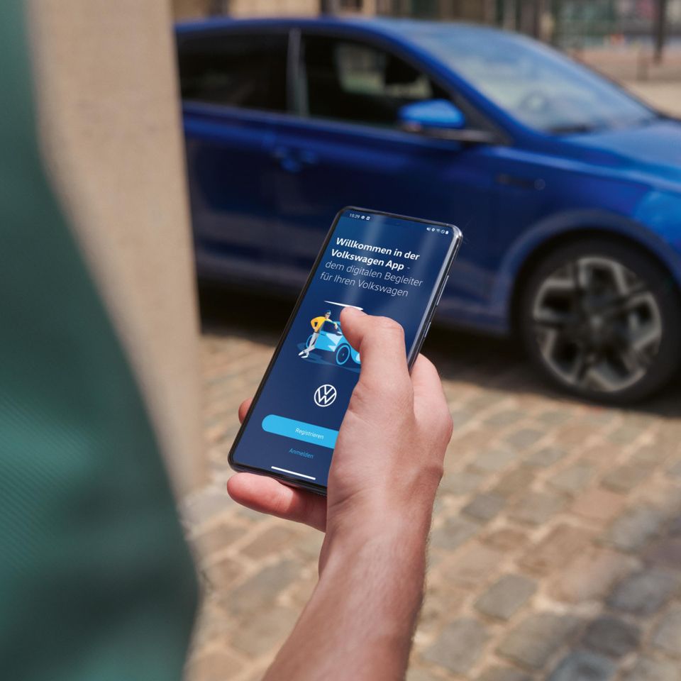  Eine Person hält ein Smartphone in der Hand mit der Volkswagen App, im Hintergrund steht ein blauer VW Polo R-Line.