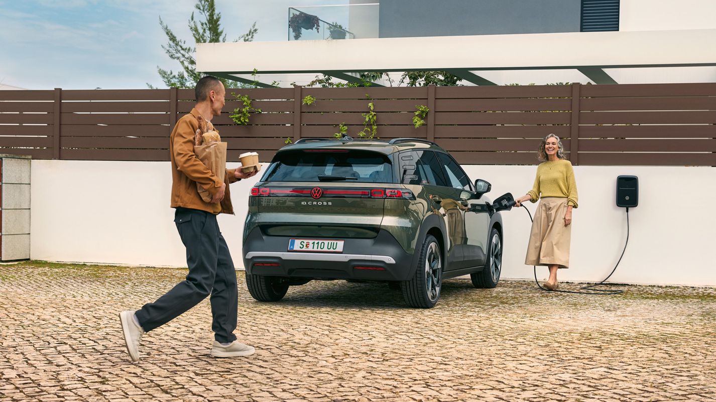 Ženska priklaplja polnilni kabel stenske polnilne postaje na zeleni VW ID. Cross, moški se ji približuje.