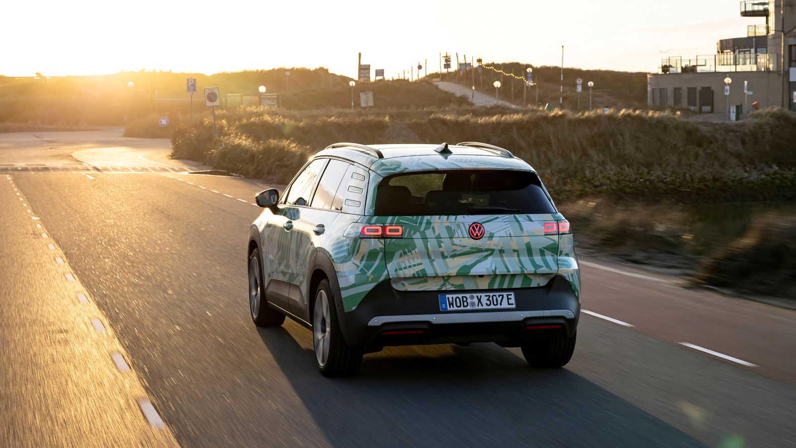 Avto vozi po cesti v pokrajini ob sončnem zahodu. Vozilo ima zeleno-belo obliko in je električni model Volkswagen SUV. V ozadju so vidne stavbe in hribi.