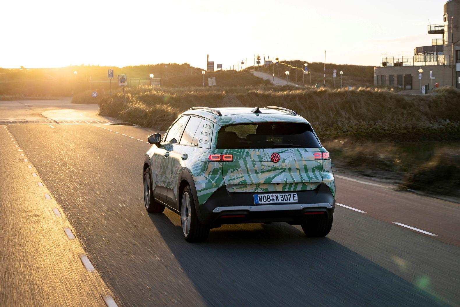 Avto vozi po cesti v pokrajini ob sončnem zahodu. Vozilo ima zeleno-belo obliko in je električni model Volkswagen SUV. V ozadju so vidne stavbe in hribi.