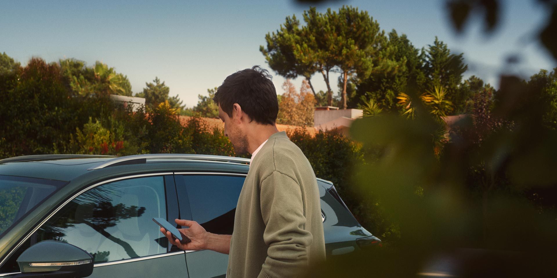 Mladenič na voznikovi strani temnozelenega VW Tiguana s pametnim telefonom upravlja opcijski sistem Park Assist Plus.