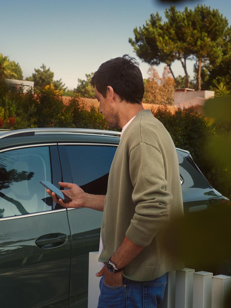Mladenič na voznikovi strani temnozelenega VW Tiguana s pametnim telefonom upravlja opcijski sistem Park Assist Plus.