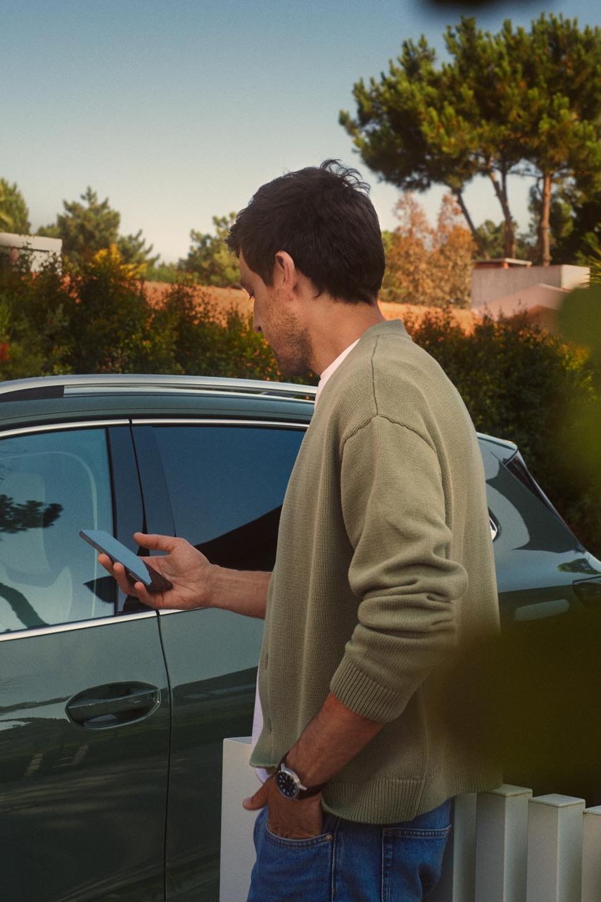 Mladenič na voznikovi strani temnozelenega VW Tiguana s pametnim telefonom upravlja opcijski sistem Park Assist Plus.