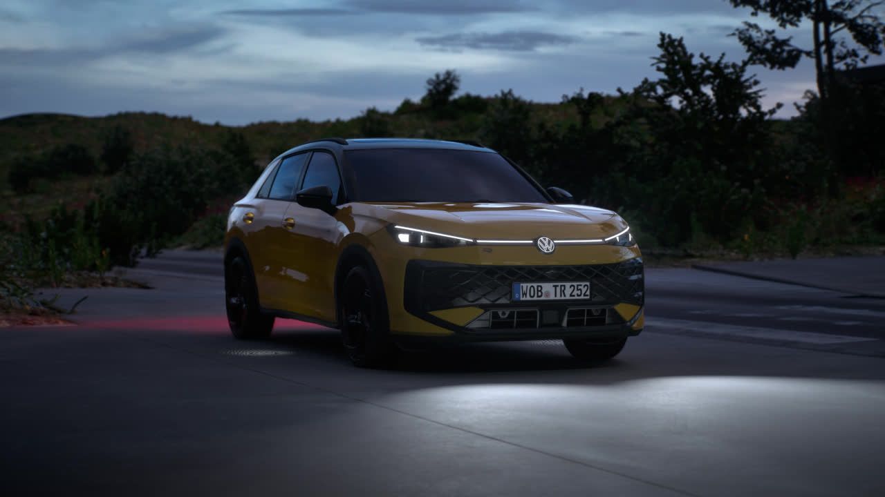 Volkswagen T-Roc rumene barve, parkiran na cesti ob večernem času. Sprednje luči avtomobila so prižgane. V ozadju so gozd in grmičevje.