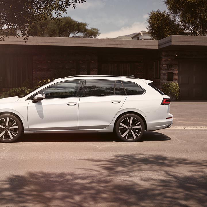 VW Golf Variant bele barve je parkiran ob mirni stanovanjski ulici pred moderno, enonadstropno hišo z veliko stekla in lesa. Avto ima velika aluminijasta platišča in športno obliko. V ospredju so senca drevesa in mehka sončna svetloba, ki ustvarjata topel, eleganten vtis.