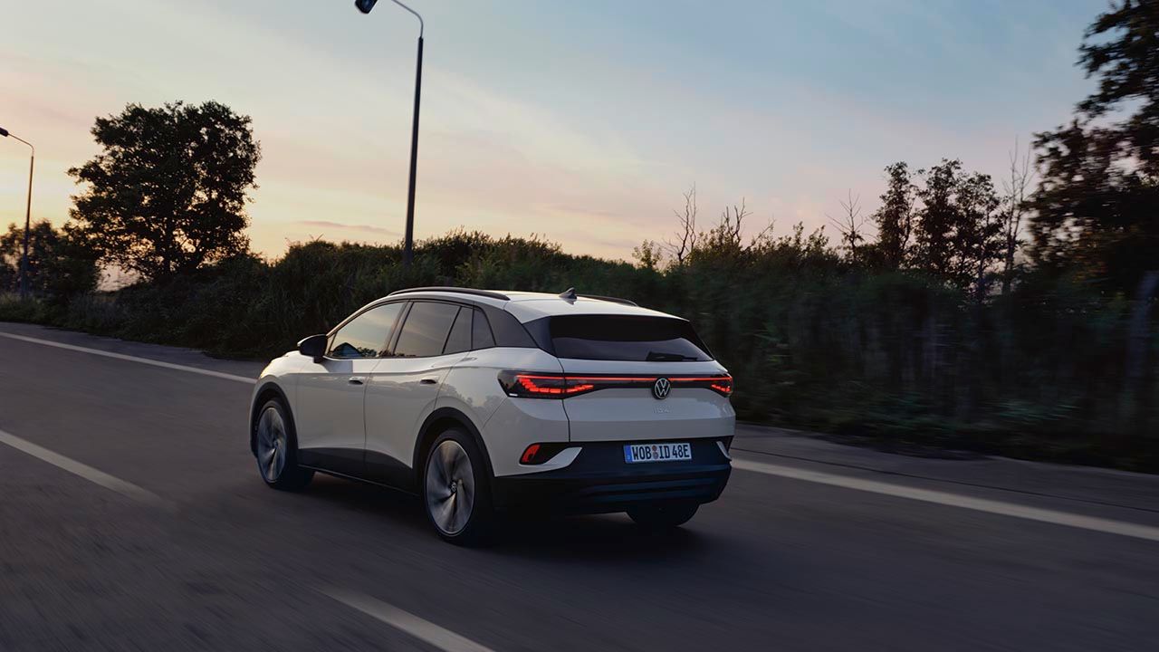 Od zadaj je prikazan bel VW ID.4, ki se ob sončnem zahodu pelje po regionalni cesti.
