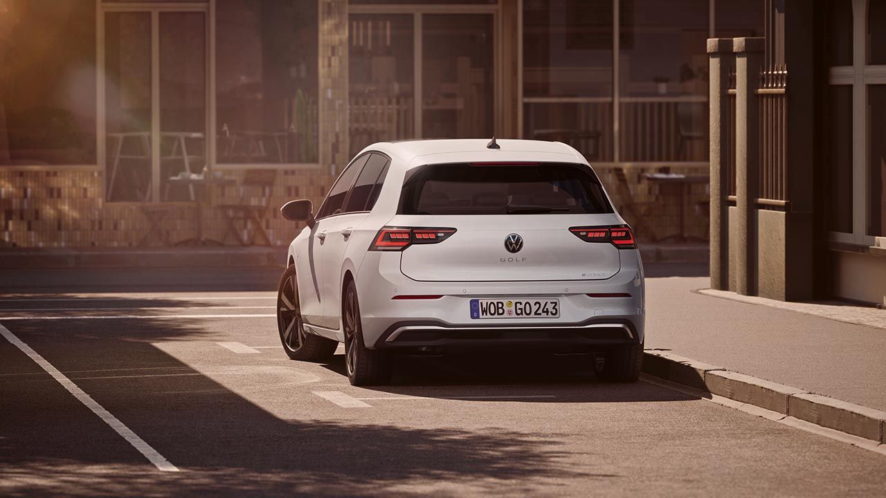 Bel avtomobil Volkswagen Golf bele barve je parkiran ob robu mirne mestne ulice. Na sliki je viden zadnji del vozila z vklopljenimi zadnjimi lučmi in registrsko tablico. V ozadju so zastekljene stavbe in mehka, topla svetloba, ki ustvarja umirjeno vzdušje.