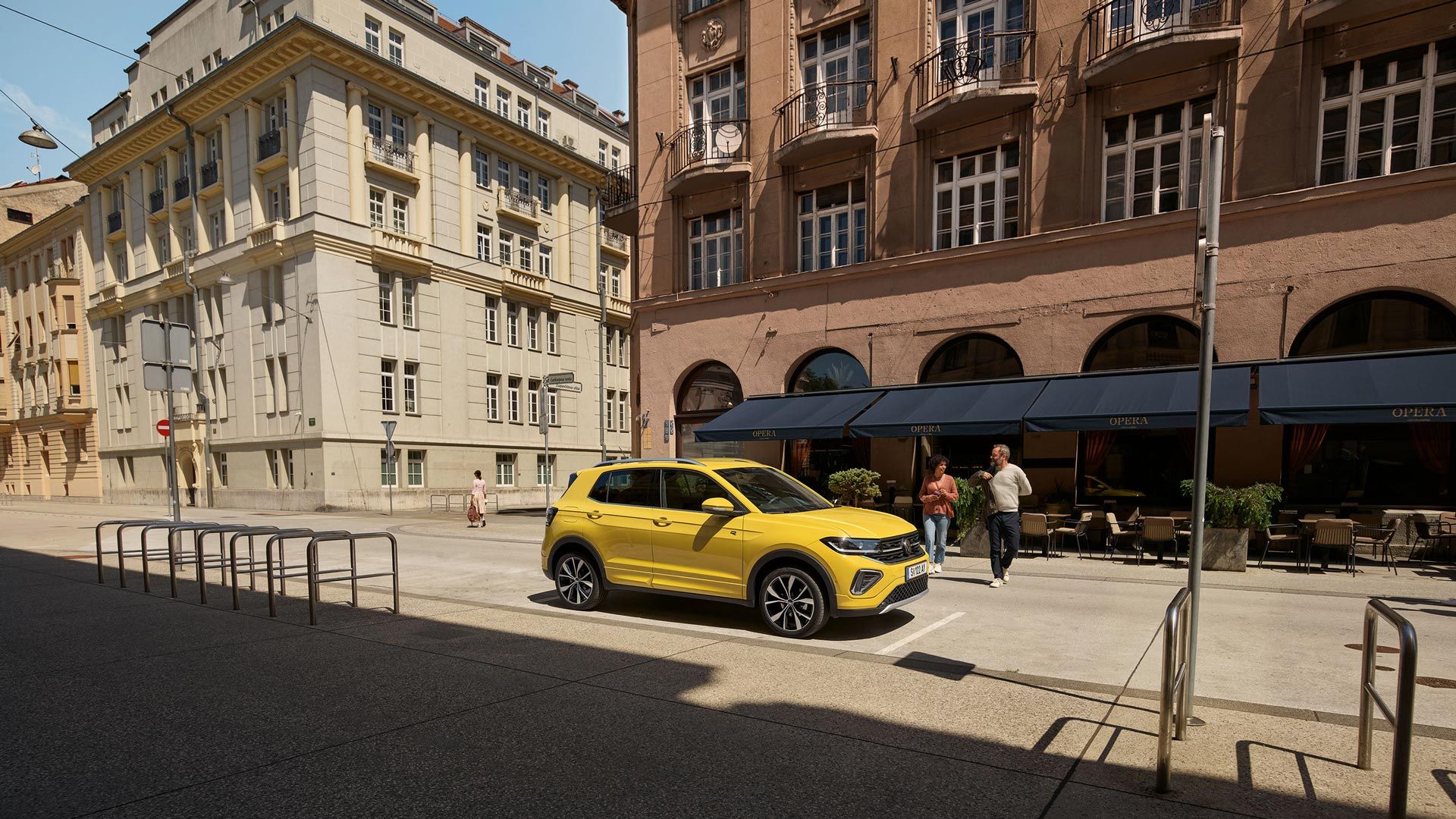 Rumen VW T-Cross je parkiran pred mestno ulično kavarno. Približuje se mu par. Avto je prikazan od strani.
